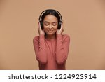 Small photo of A young girl mixedrace listens to her favorite music in big black headphones, closing her eyes from pleasure, enjoyment, music lover, likes lyric songs about love, on beige background