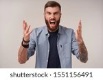 Small photo of Wroth young bearded brunette male with trendy haircut shouting angrily and raising palms emotionally, frowning eyebrows with wide mouth opened while looking at camera, isolated over white background