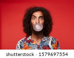 Amazed pretty brown-eyed bearded guy with dark curly hair making bubble with gum and looking surprisedly to camera, standing over red background in shirt with floral print