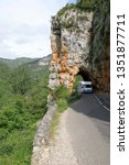 Small photo of Tarn Gorge France circa May 2016. Large motorhome driving gingerly through a narrow natural arch along a narrow road with long drop down gorge to its right. Verdant lined banks, White cloud.