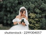 Small photo of Brunette Bride in a voluminous white dress with open shoulders and a long veil, smiling, posing with a bouquet of white roses. Portrait of the bride. Beautiful makeup and hair.