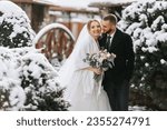 Small photo of Beautiful and happy bride and groom are having fun among snowy trees. Bride and groom in the winter park. Bride with a bouquet of flowers in a wedding dress and poncho. Groom in a black coat.