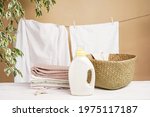 White linen is dried on a clothesline against the background of a beige wall with a houseplant. A stack of colored laundry, a laundry basket, bottles of detergent and fabric softener are on the table.