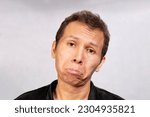 Small photo of a close up of a sad and funny Asian man looks to be upset unhappy and chapfallen , looking at camera , in black shirt on gray background