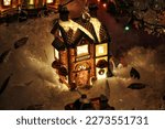 An aerial view of a festive Christmas village with quaint houses and street lights adorned with decorative lights, covered in a blanket of soft snow