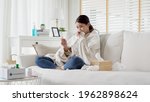 Small photo of Sick young asian woman sit under blanket on sofa and sneeze with tissue paper at home. Female blow nose, coughing, sneezing, check temp in tissue at home, suffering from flu. Cold and fever concept