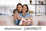 Small photo of Happy time Mother day grown up child looking at camera cuddle hug give flower gift box red heart card to mature mum. Love kiss care mom asia middle age adult people smile enjoy sitting at home sofa.