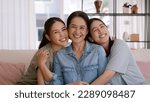 Small photo of Happy good warm time Mother day two grown up kid child looking at camera cuddle hug arm around mature mum. Love kiss care mom asia middle age adult three people smile enjoy sitting easy at home sofa