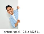 Portrait of happy young Asian man in casual clothes hiding behind blank white billboard for mockup template isolated on white background