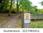 Shallow focus of a laminated Dogs On Lead sign attached to a wooden fence at the entrance to a nature reserve. A woman and her dog can be seen.