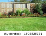 Shrubs And Flowers In A Border Surrounded By A Wooden Panel Fence And Grass Lawn In A Back Garden.