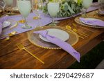 The elegant wooden table is decorated with gold linens and bright metallic accents in gold, green, and purple colors, topped with purple napkins and plates in a yellow and purple color scheme. The tab