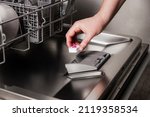 Close-up of female hand inserting dishwasher tablet into open automatic stainless built-in dishwasher machine with dirty utensils inside in modern home kitchen. Household, housekeeping domestic life