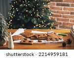 Homemade gingerbread house building blocks with glaze laying on table with decorations, lanterns in living room decorated with defocused New Year lights, Christmas fir tree. Holiday mood for kids