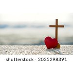 Red heart with wooden Christian cross on gravel floor in morning light, beach sea as background. Jesus love you. Faith hope believe in God. Believe in salvation. Christianity background concept.