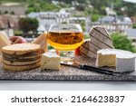 Small photo of Cow cheeses of Normandy region - camembert, livarot, neufchatel, pont l'eveque and glass of apple cider drink with houses of Etretat village on background, Normandy, France