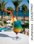 Colourful frozen Rainbow Paradise cocktail drink served in glass at pool bar overlooking blue pool, sea and palm trees, relax and holidays at sea