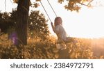 Small photo of Blonde little girl swings back and forth on wooden swing in sunny summer park and gentle breeze ruffles light hair. Blonde little girl swings in sunny park with big trees and high grass on meadow