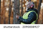Small photo of Elderly african american forestry engineer in noiseisolating headphones in forest old mature foreman forester thinks over work plan for cutting trees by entering data into tablet evaluating situation