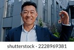 Small photo of Happy smiling Asian adult 40s man holding keys portrait outdoors win reaction victory celebrate male realtor real estate agent investor buyer selling property new building office dwelling skyscrapers