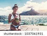 Small photo of Portrait of serious hottie Brazilian girl sitting on the embankment bench and holding fresh opened coconut for drinking; with cityscape, bay, and Rio de Janeiro mountains the Two Brothers behind her