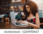 Small photo of Cute curly hipster female freelancer in a street bar with spritz cocktail working on her netbook; drop-dead gorgeous caucasian female using her laptop while sitting in an outdoor cafe with beverage