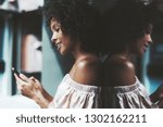 Small photo of A drop-dead gorgeous African-American girl with a medium curly Afro hair outdoors is leaning against a glass wall which is totally reflecting her while messaging a friend via the smartphone