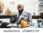 Small photo of Portrait of a baldheaded adult black dandy guy with a well-groomed beard, in a custom made elegant costume and eyeglasses sitting in a street cafe with a cup of delicious coffee and reading a magazine