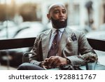 Small photo of Portrait of a dapper mature hairless black man entrepreneur with a nice beard, in a custom elegant grey costume, sitting in an outdoor cafe on a rainy day and pensively looking into the distance