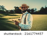 Small photo of A portrait of a dashing mature bearded black man in a fashionable outfit with a straw hat and braces standing on a golf field in the evening with a sunset, holding a golf club and a cigar in the hands