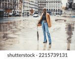 Small photo of A young svelte black female with short hair painted white is standing on the wet ground of Lisbon city center after the rain and leaning on her umbrella, with a copy space area on the left