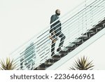Small photo of A bald black bearded man walks up the stairs. The African male is ascending a white metallic staircase on a shiny day, wears a dark suit and holds a leather purse in a very clean and cactus aesthetic