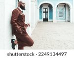 Small photo of A black man with an unshaven beard wearing a Bordeaux suit leaned against a white stone pillar. Side view of a dandy male holding a pair of eyeglasses looking dapper in a fancy suit adjacent to a wall
