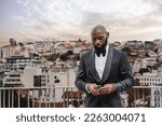 Small photo of Portrait of a charming and gallant black man, well dressed in a black suit, a bow tie, and a white shirt, this one in a closed semblance looking at his smartphone on a terrace overlooking the city