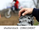 Small photo of Dirt bike race racing. Extreme sports. Motocross. Man's hand on old knubby bumpy tire. Spectator watching races. Selective focus. Blurred background man riding dirtbike. Competition. Track. Course.