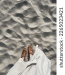 Small photo of Women feet on desert beach sand. Aesthetic summer vacation background. Woman in white dress and leather sandals on the dune sand. Top view