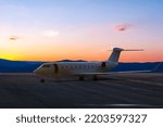 Small photo of Modern white private jet with an opened gangway at the airport apron on the background of a picturesque sunset