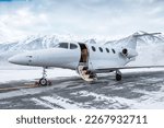 Small photo of Modern white executive jet plane with an opened gangway door at the winter airport apron on the background of high scenic snow capped mountains
