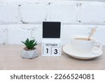 Wooden calendar with date October 13 and plant with coffee cup on table against brick wall background. Deadline, planning, business concept