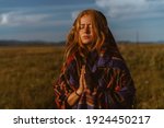Small photo of in the steppe against the sky stands folding prayerfully the hands of a girl in an ethno cape with red hair covering her eyes with an expression of entreaty on her face. High quality photo