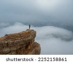 Soft focus. Tourist (woman) stands on the dangerous cliff edge of plateau on high altitude under snowy cloudy sky in foggy morning. Person standing above the clouds.