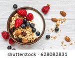 bowl of oat granola with yogurt, fresh raspberries, blueberries and nuts on white wooden board for healthy breakfast. top view