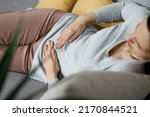 Small photo of Sick young woman suffering from menstrual pain. Woman with hands squeezing belly having painful stomach ache or period cramps sitting on sofa, Abdominal pain, gastritis, and painful periods concept