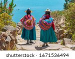 Two indigenous Quechua women in traditional clothes walking down the path to the harbor of Isla Taquile (Taquile Island) with the Titicaca Lake in the background, Peru.