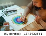 Young woman colouring mandala with markers and white rosary on table with cup of coffee near flower vase at home. Art therapy and meditation concept. Hobby and leisure during quarantine. Wellbeing.