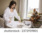 Small photo of Smiling young plus size woman folding linen nightdress in PVC container and metal basket storage organizing. Beautiful female sorting organic cotton textile on table comfortable housekeeping