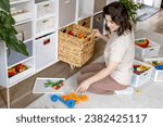 Small photo of Smiling female housewife childish toys storage organizing sorting details with basket and PVC container. Woman cleaning kids room filling cabinet by multicolored playthings comfortable maintaining