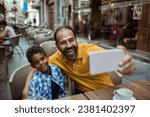 Small photo of Young stepfather taking selfies with his stepson in a outdoor cafe sitting area in the city