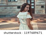 City secrets. Beautiful smiling woman wearing a light dress looking away over the shoulder while walking outdoors. She happily turned back to well known masculine vice. City architecture.