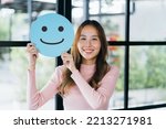 Small photo of Asian woman holding blue happy smile face on paper cut, user giving good feedback rating, think positive , customer review, assessment, of mental health day, Compliment Day, satisfaction concept.